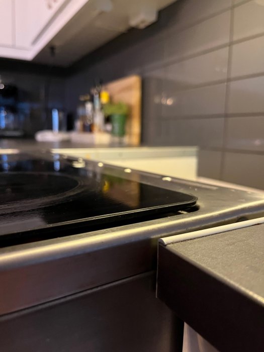 Glaskeramikhäll i ett kök, närbild, oskarp bakgrund, vita luckor, grått kakel, träskärbräda, bänkskiva, hushållsflaskor.