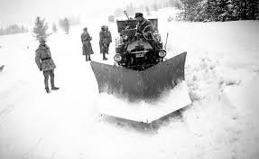 Personer i vinterlandskap, snöplogfordon, vitt landskap, tjock snö, kallt väder, arbetsklädda individer, gråskaligt foto.