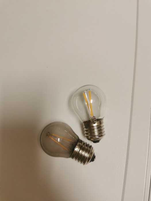 Två glödlampor med synliga filament, olik storlek, vit bakgrund, ej tända, en står, en ligger.