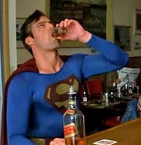 En person klädd som Superman dricker från en shotglas på en bar, ser missnöjd ut, whiskeyflaska framför honom.