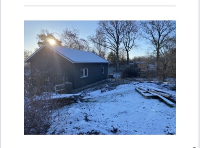 Vinterlandskap med hus, snö, träd, soluppgång, vedstaplar, byggmaterial och en klar himmel.