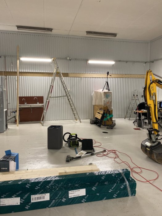 Industriell verksted med byggeprosjekt, stiger, trevirke på gulvet, og en liten gravemaskin. Veggene har liggende treverk for å støtte et nytt loft.