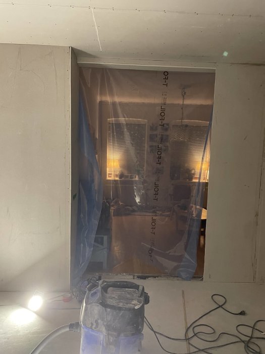 Byggarbetsplats med plasttäckt öppning, gipsskivor, dammsugare, arbetslampa, ofärdiga väggar och tak, rörande inomhusrenovering.