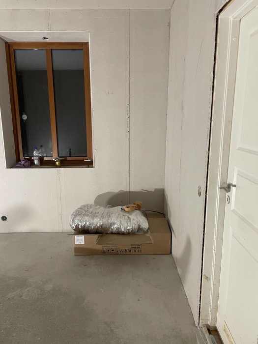 Ett ofärdigt rum med gipsskivor, kartong, fönster, vit dörr och en silvrig kudde.