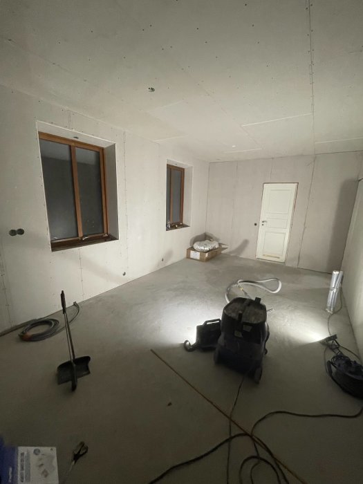 Omfattande renovering pågår, oslipat golv, osprayade väggar, fönster, dörr, byggdammsugare, byggbelysning, renoveringsverktyg.