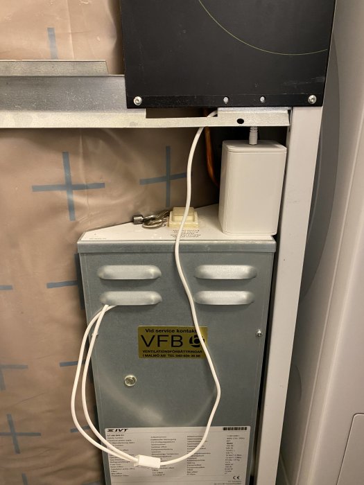 En teknisk installation med kablar, ventilationsbox, etikett för servicekontakt och telefonnummer.