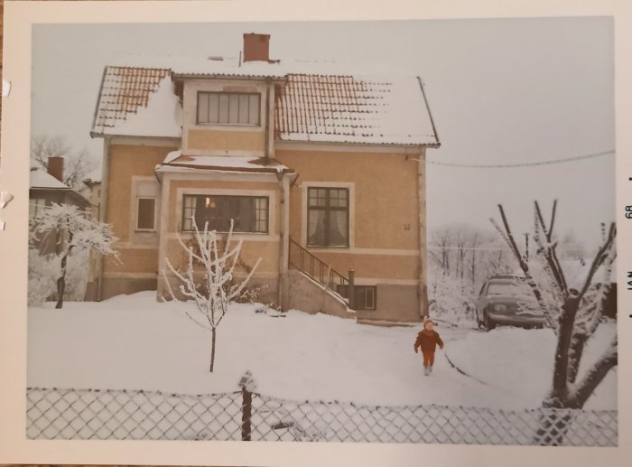 Ett gult hus i snölandskap, med barträd, staket, och barn framför.