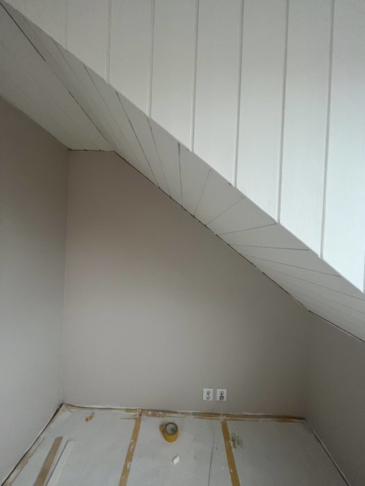 En tom hörna i ett rum under renovering, med snedtak och vita väggpaneler.
