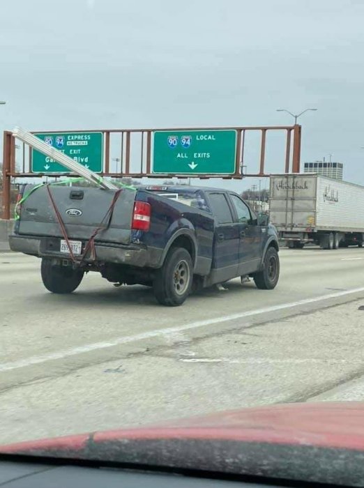 Pickup med trasig baksida, provisoriskt reparerad med rep, kör på motorväg. Vägskyltar ovanför.