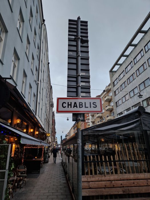 Stadsgata med högt torn av tomma affischramar, skylt "Chablis", uteservering, och en person går.