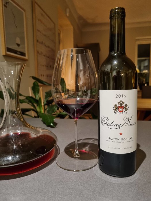 Ett glas rödvin, dekanteringskanna, vinflaska mot en duk, inomhusbelysning, hemmiljö, Chateau Musar 2016.