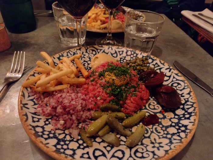 Tallrik med tartar, pommes, inlagda grönsaker, rödbetor. Mönstrad tallrik. Vin och vattenglas i bakgrunden.