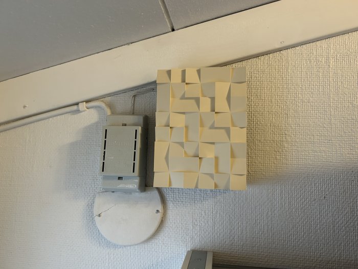 Väggmonterad termostat, gula post-it-lappar, kabel, vit texturerad vägg, grått tak, delvis doldt synfält.