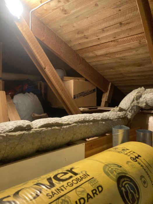 Ett oupplyst vindsutrymme med synliga träbjälkar, isoleringsmaterial, förvaringslådor och en tänd lampa.