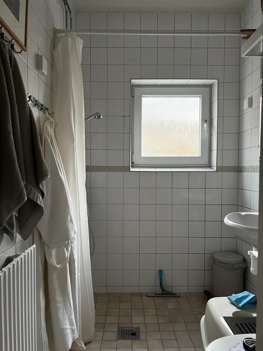 Ett enkelt badrum med duschdraperi, toalett och handfat, kakelväggar, fönster och städutrustning.
