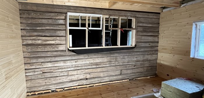 Inomhusrenovering, råtimrade väggar, öppet fönster, kontrast mellan obehandlat och behandlat trä, renoveringsmaterial synliga.