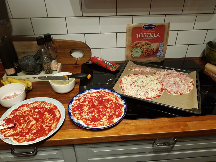 Förberedelse av pizza med tomatsås, skinka, ost; tortillapaket; köksverktyg och ingredienser på köksbänk.