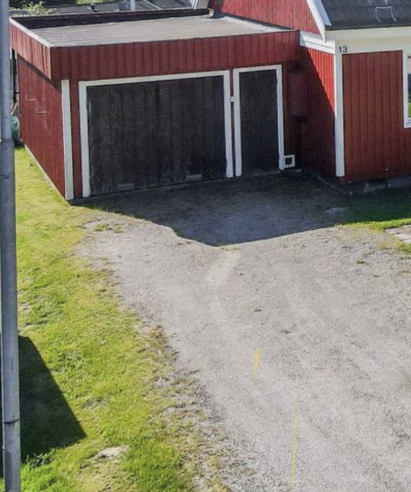 Röd träbyggnad med två garageportar, grå grusdrivbana, grönt gräs, soligt, del av husnummer 13 synligt.