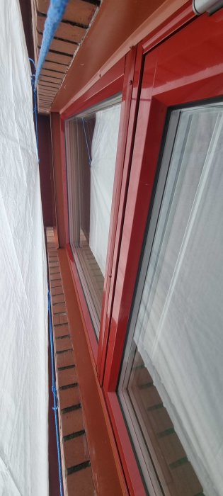 Mellan husvägg och fönster, rött fönsterkarm, tegel, snöre, presenning, snedvinkel, reflektion i glas.