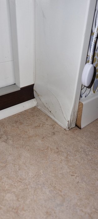 Skadad dörrfoder i hörnet, vitmålad vägg, beige matta, träkloss som dörrstopp, lägenhetsinteriör med slitage.