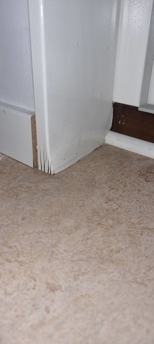 Närbild på vit dörrkarm och golv, med skador och slitage synliga.