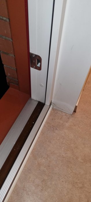 Halvöppen dörr till ett rum, vit dörrkarm, tegelvägg, golvtröskel, matta, och smutsiga spår.
