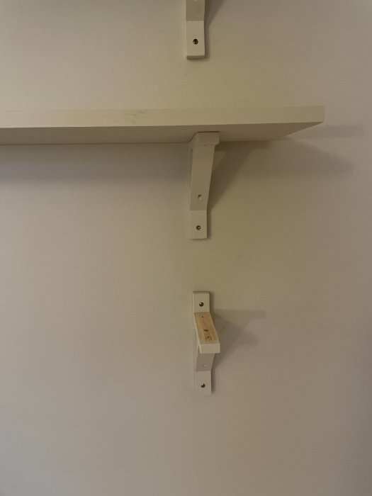 Två vita hyllor på en vägg, nedre saknar sin hyllskiva, synliga fästen och skruvar.