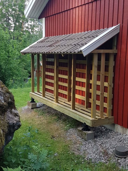 Röd stuga, vedstapel, täckt vedförvaring, grönt gräs, grå plattor, bebyggd natur, träd, byggnadsstöd, dagtid.