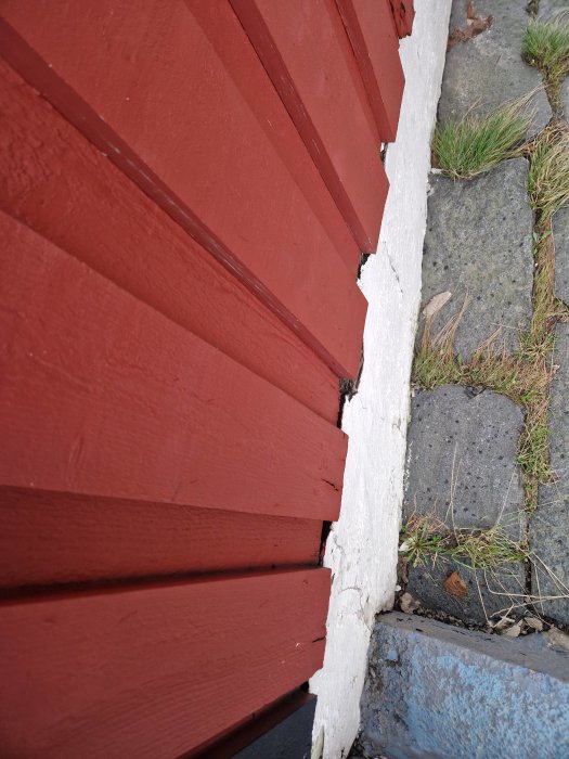 Röd träpanel möter vit mur, grästuvor och stenplattor på marken. Detaljbild av hus och trottoar.