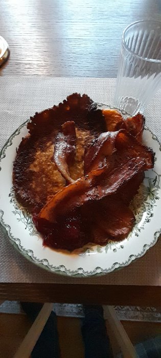 Pannkaka och bacon på tallrik, klart för måltid, enkelt träbord, genomskinligt glas, hemmiljö.