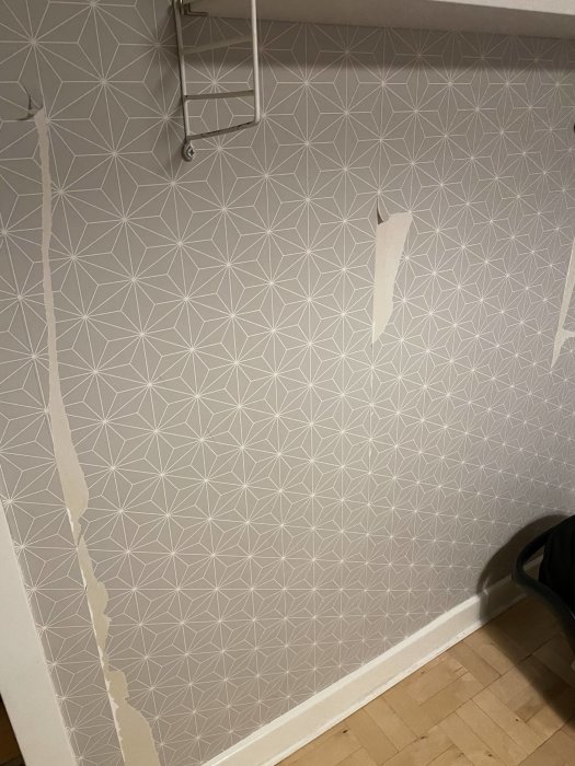 Skadad geometriskt mönstrad tapet, vit vägg, trägolv, delvis avskalad, hängande hyllsystem överst.