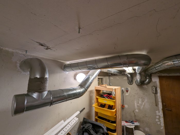 Ett rörigt källarrum med synliga ventilationstrummor i taket och verktygshylla.
