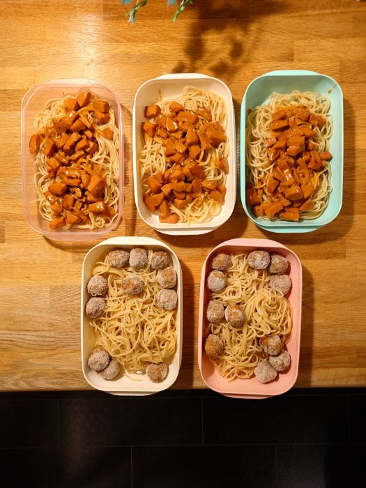 Fyra matlådor med pasta, två med köttbullar, två med tofu, på träbord.