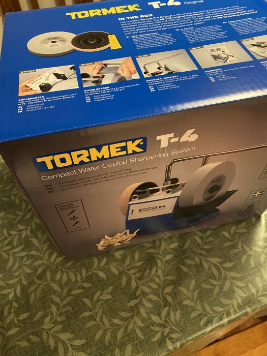 En Tormek T-4 slipmaskins förpackning på ett blommigt bord. Verktygsslipningssystem, instruktionsbilder, flerspråkig text.