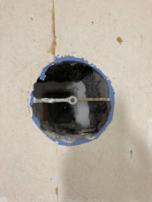 Rund väggöppning med synliga rör och kablar, omgiven av blå målartape och gipsskivor.