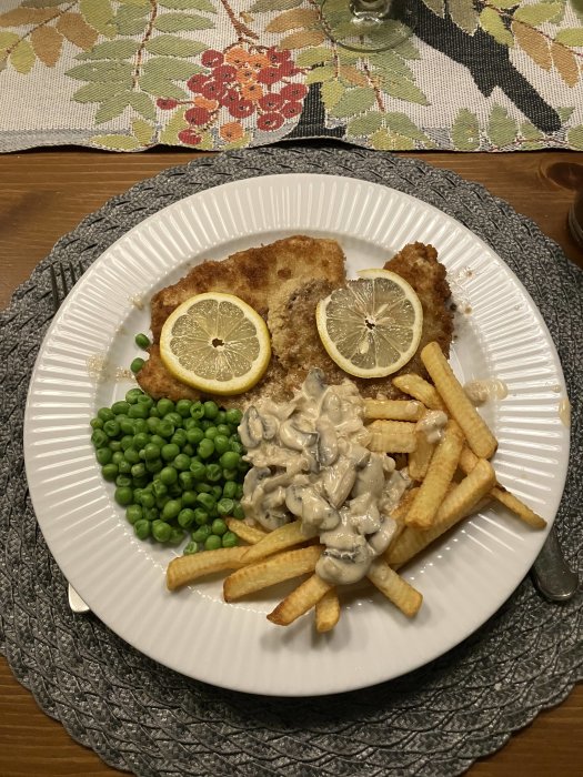 Middagstallrik med schnitzel, citronskivor, gröna ärtor, pommes frites och svampsås på ett dekorerat bord.