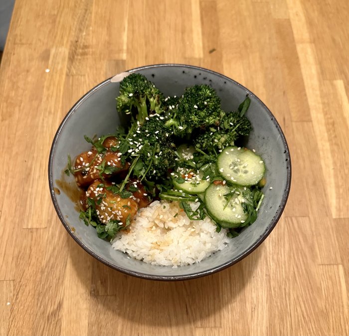 Maträtt i skål: ris, broccoli, agurk, teriyakikyckling, sesamfrön, på träbord.