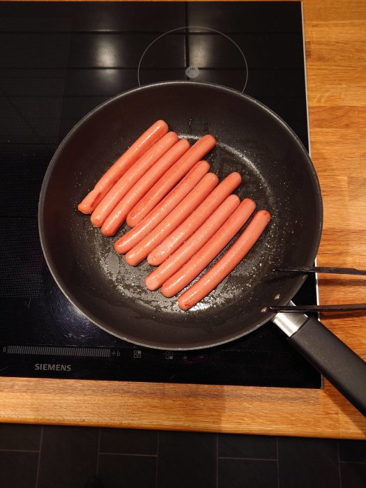 En stekpanna på spishäll med stekande korvar ovanpå en träbänk.