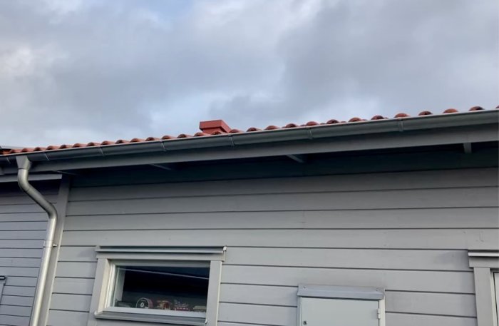 Grått hus, röda takpannor, skorsten, öppet fönster, stuprör, molnig himmel, bostadsarkitektur, dagtid.