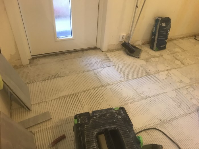 Ett rum under renovering med kakellim på golv, arbetsverktyg och oavslutad installation av golvplattor.