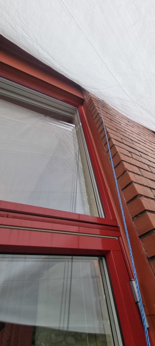 Röd fönsterram, tegelvägg, vitt tygtak, blått rep, utomhus, delvis öppet fönster, arkitektoniska detaljer.