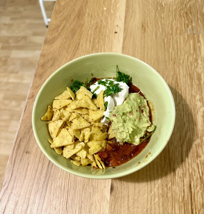 En skål med nachos, guacamole, gräddfil, chili, gröna örter på ett träbord.