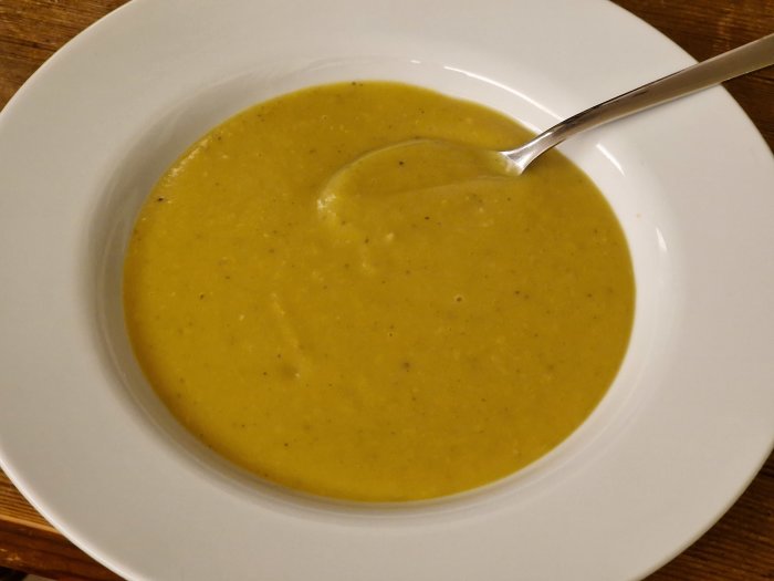 En djup tallrik med krämig, gul soppa och en sked, på ett träbord.
