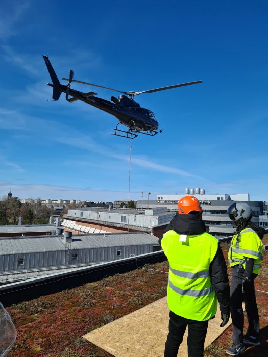 Helikopter lyfter last, två personer i säkerhetsvästar och hjälmar observerar, stadsmiljö i bakgrunden, klarblå himmel.