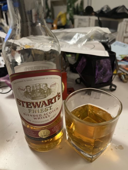 Flaska av Stewart's Finest Scotch Whisky och ett glas med whisky på ett rörigt skrivbord.