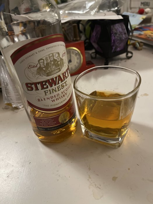 Flaska med Scotch whisky och fylld whiskyglas på rörigt köksbord.