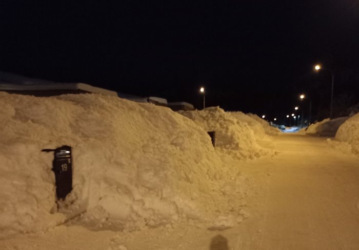 Gata med snöväggar nattetid, gatubelysning, brevlåda synlig, snötyngda tak, kallt vinterlandskap.