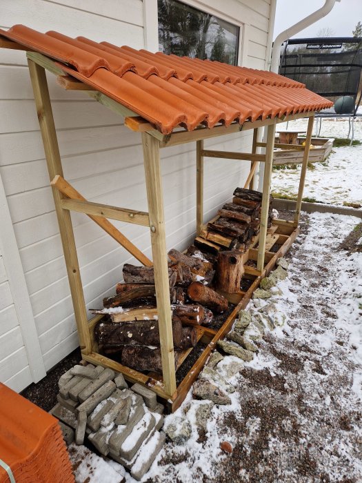 En vedbod med tegeltak, staplad ved, snötäckt mark, och stenplattor vid husvägg.