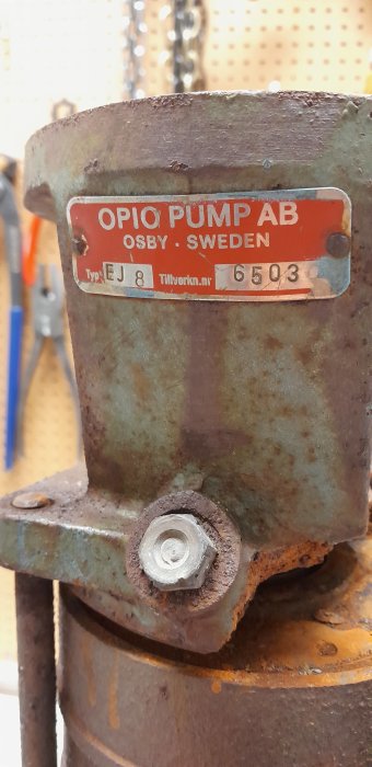 Rostig, industriell pumpdel med etikett "OPIO PUMP AB, OSBY·SWEDEN", fästelement och bakgrund med verktyg.