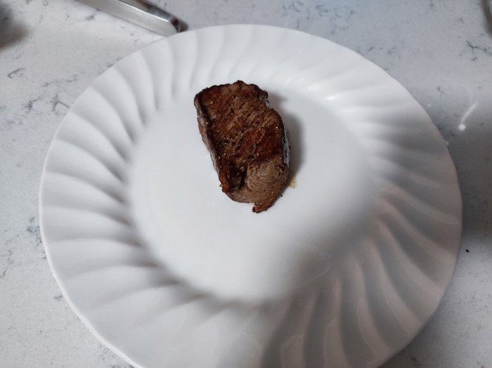 Ett grillat köttstycke på en vit tallrik med vågig kant, på ett marmorbord.